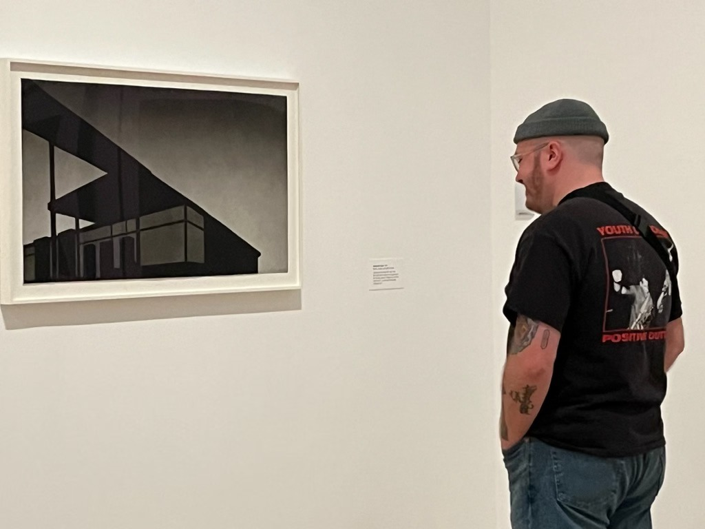 James Coleman Howes looking at an artwork in a gallery setting, wearing a beanie and a "Youth of Positive Out" t-shirt.